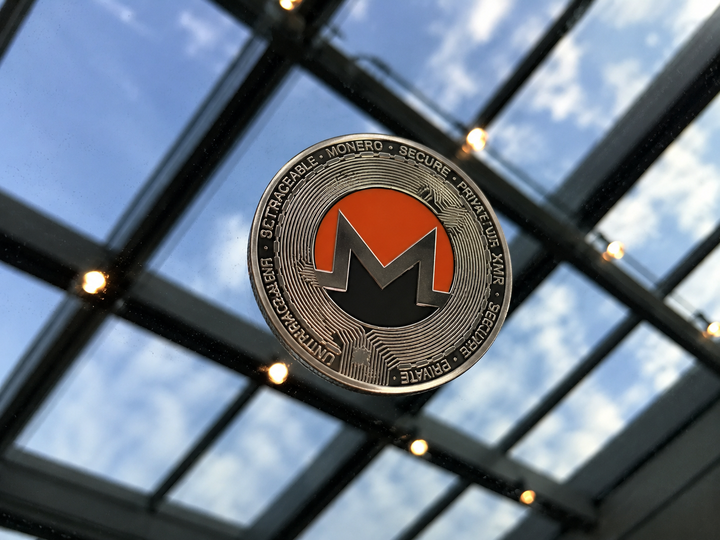 Ein silberner Monero-Token mit dem orangefarbenen XMR-Logo liegt auf einer spiegelnden Glasoberfläche unter einem Oberlicht.