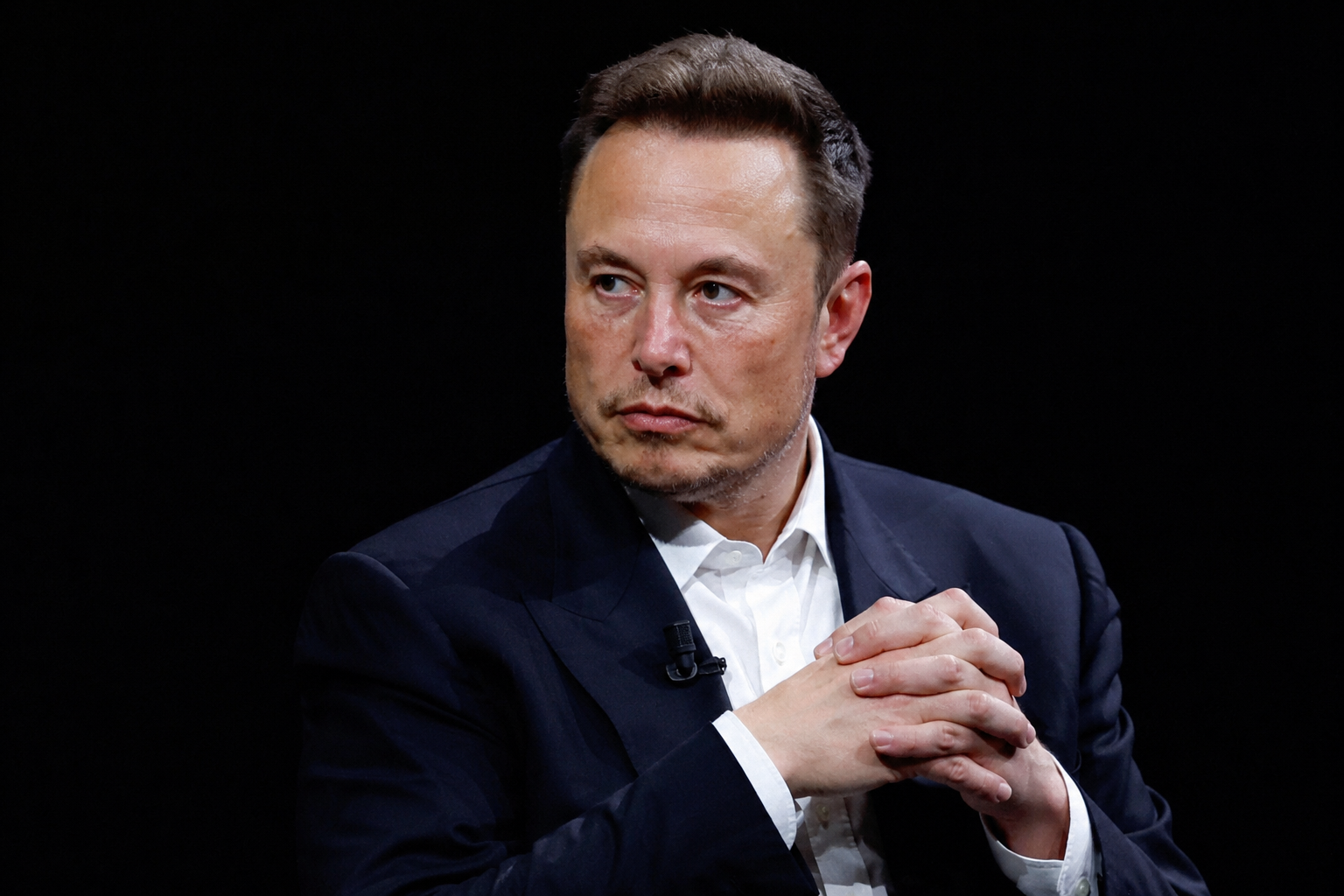 Elon Musk sits with clasped hands while attending a public technology event.